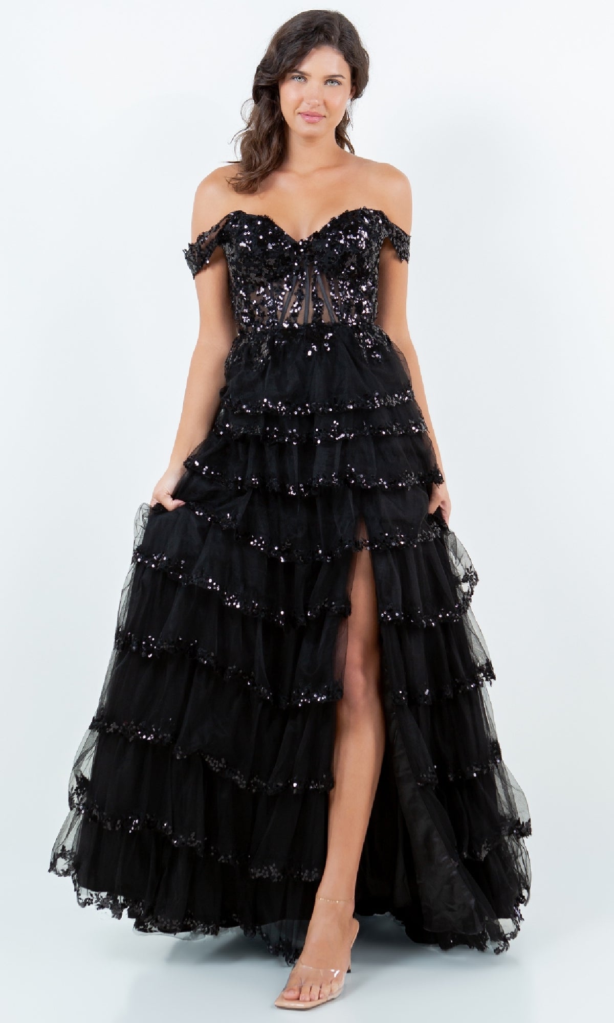 A woman models the Ruffle Prom Ball Gown: Cinderella Couture 8228J, an off-the-shoulder black tiered tulle dress with sequin embroidery and a thigh-high slit, standing against a plain white background.